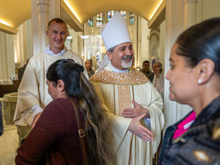 Bishop in white vestments greets worshippers with a handshake inside a bright church, smiling warmly.