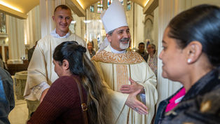 Bishop in white vestments greets worshippers with a handshake inside a bright church, smiling warmly.