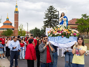 People carry a flower-covered Virgin Mary statue in a church procession, with a crowd and cathedral towers under a cloudy sky.