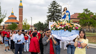 People carry a flower-covered Virgin Mary statue in a church procession, with a crowd and cathedral towers under a cloudy sky.