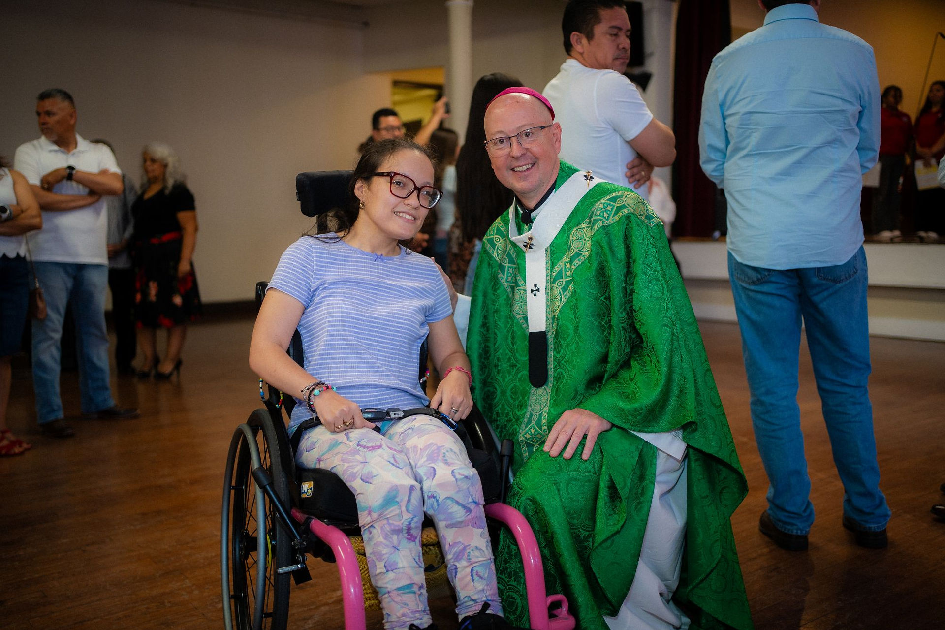 Smiling woman in a wheelchair poses with a bishop in green robes at an indoor gathering, with people standing in back.