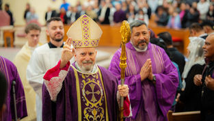 A bishop in ornate purple robes and a mitre raises his hand in blessing, surrounded by clergy in a church filled with people.