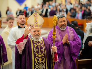 A bishop in ornate purple robes and a mitre raises his hand in blessing, surrounded by clergy in a church filled with people.