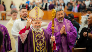 A bishop in ornate purple robes and a mitre raises his hand in blessing, surrounded by clergy in a church filled with people.