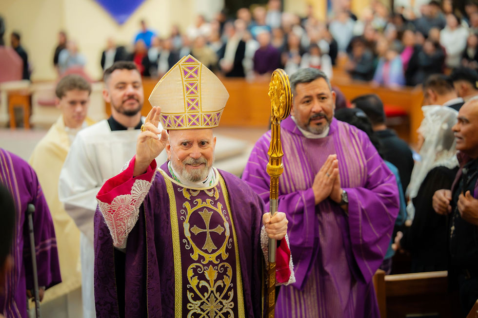 A bishop in ornate purple robes and a mitre raises his hand in blessing, surrounded by clergy in a church filled with people.