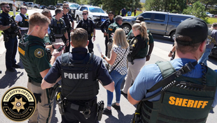 Police and sheriffs gather on a street, engaged in discussion. Vehicles and trees are in the background. Officers wear uniforms with visible badges.