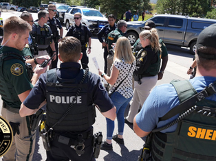 Police and sheriffs gather on a street, engaged in discussion. Vehicles and trees are in the background. Officers wear uniforms with visible badges.