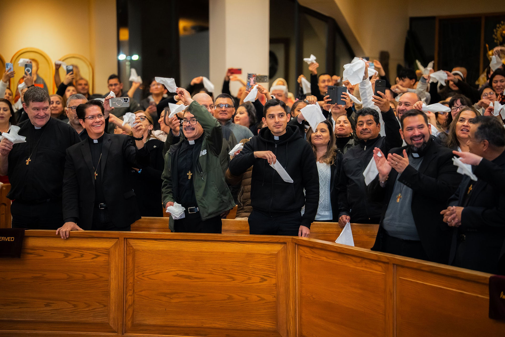 A joyous crowd in a church waves white cloths and takes photos. People are smiling, wearing dark clothes, with wooden pews in the foreground.