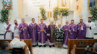 Clergy in purple vestments pose with a woman beside an altar adorned with flowers and religious artwork. Text on wall reads "Aquí que soy tu madre".
