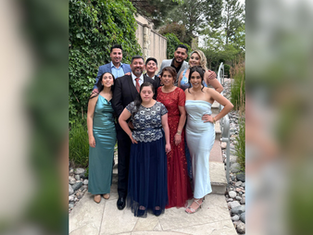 A group of nine people in formal attire pose happily outdoors, surrounded by greenery. The mood is joyful and celebratory.