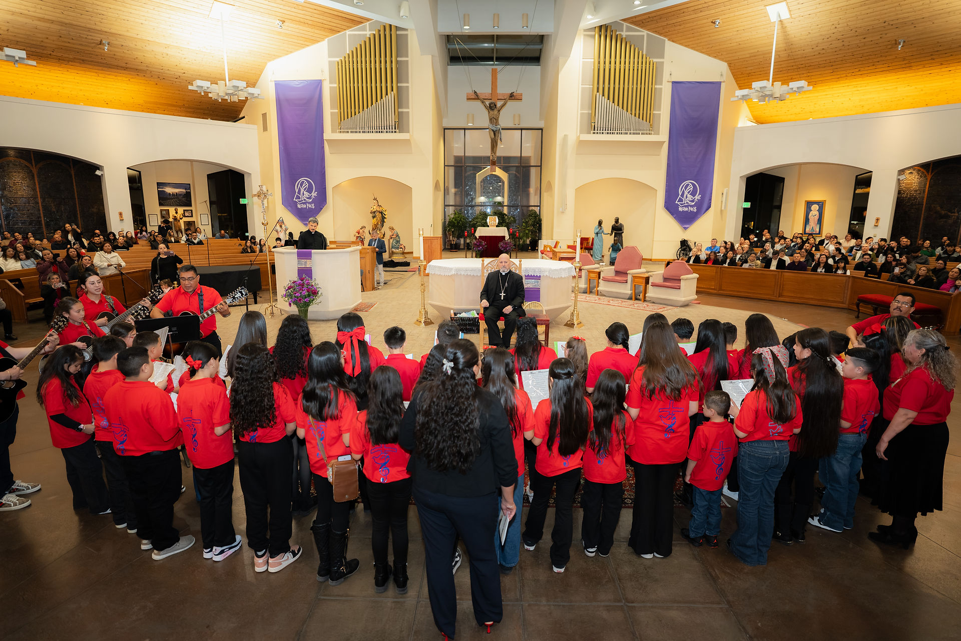 A man in black robes sits on a chair in a church, facing children in red shirts. Background includes altar, candles, and statues.