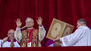 Clergy in ornate robes stand before a red curtain. One man raises hands in blessing, flanked by assistants holding an elaborate book.