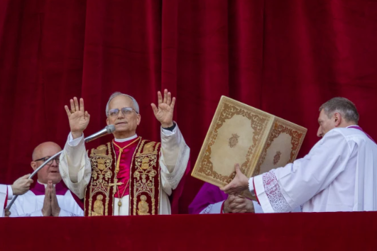 Clergy in ornate robes stand before a red curtain. One man raises hands in blessing, flanked by assistants holding an elaborate book.