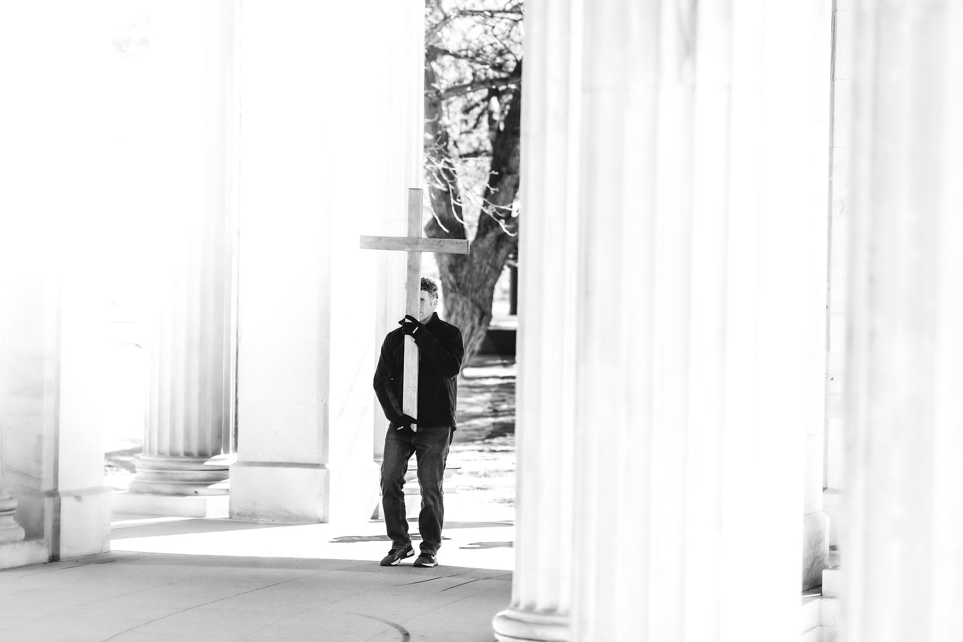 A man carries a wooden cross between tall columns in a black-and-white image during a prayer procession.