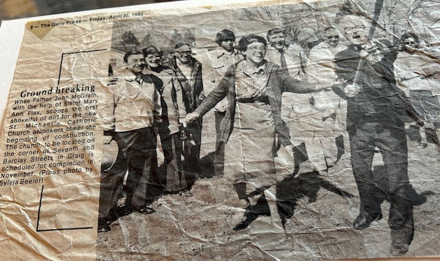 People gather at a groundbreaking ceremony, smiling and holding shovels. The setting is outdoors; a newspaper clipping date is visible.