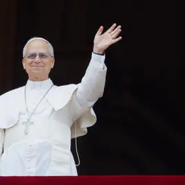 Pope in white robes smiles and waves from a balcony against a dark background.