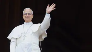 A man in white religious attire waves from a balcony. He wears glasses and a cross necklace, with a dark background and a red railing.