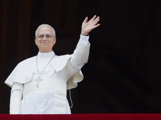 Pope in white robes smiles and waves from a balcony against a dark background.