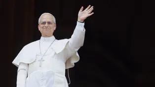 Pope in white robes smiles and waves from a balcony against a dark background.