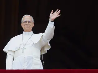 Man in white papal attire waves from a balcony against a dark background. He looks content, and a cross pendant is visible.