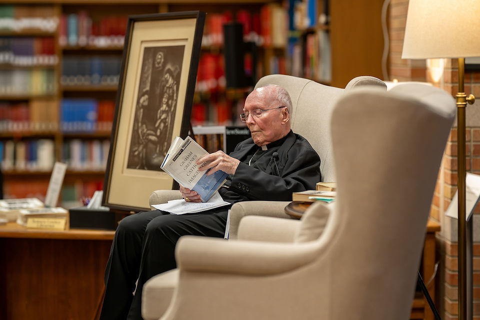 Elderly man reading a book titled "Catholicism" in a cozy library, seated in an armchair. Framed artwork and warm lighting in background.