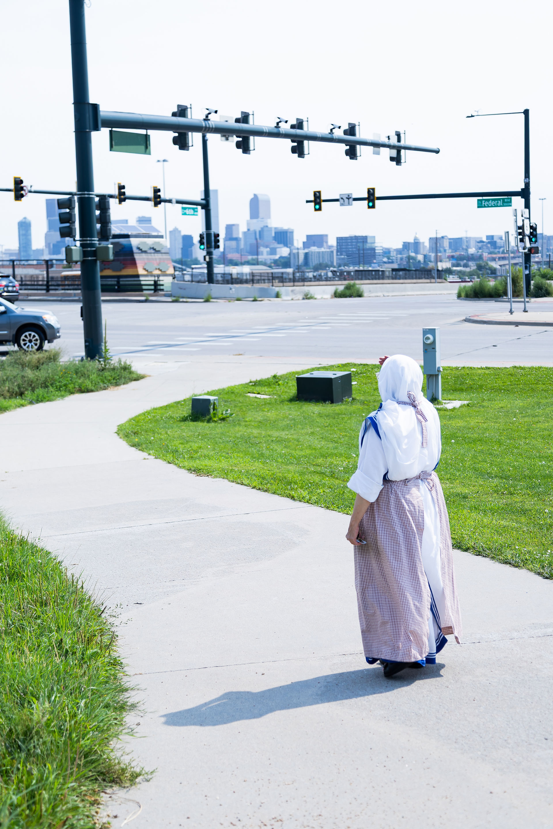 A person in a religious habit walks on a sidewalk towards traffic lights, against a city skyline backdrop. Signs read "Federal Blvd."