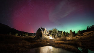 Stone chapel illuminated at night, surrounded by trees and grass. Aurora borealis in sky with red and green hues. Reflective water in foreground.