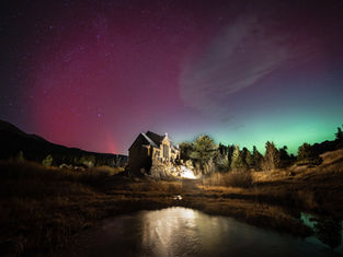 Stone chapel illuminated at night, surrounded by trees and grass. Aurora borealis in sky with red and green hues. Reflective water in foreground.