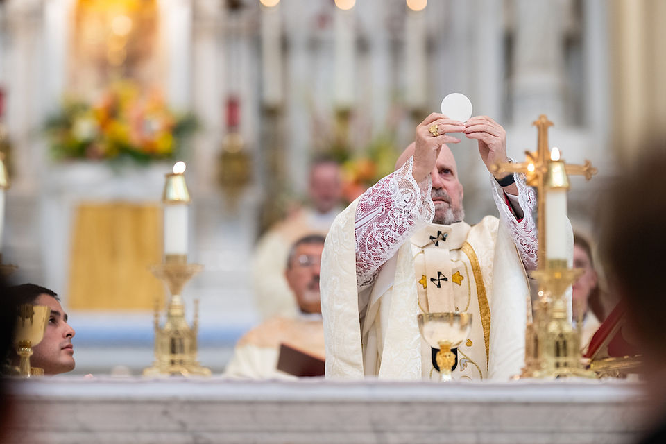 Bishop in ornate vestments holds up a Host during a service, surrounded by candles and blurred altar, in a reverent church setting.