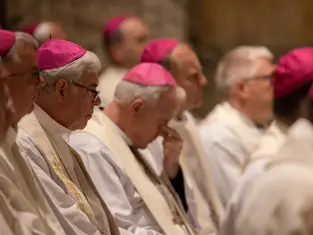 Clergy in white robes and pink caps sit solemnly in a dimly lit church; one man bows his head in prayer.