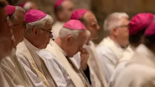 Clergy in white robes and pink caps sit solemnly in a dimly lit church; one man bows his head in prayer.