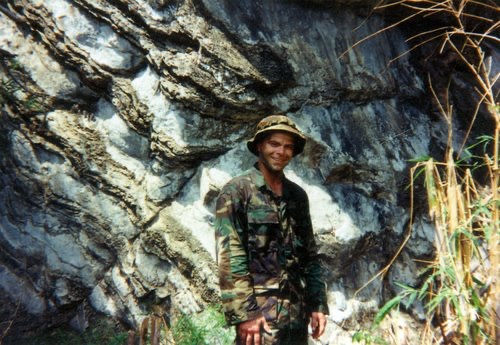 Person in camouflage gear smiles near rocky cliff, with tall grass nearby. Sunlight creates shadow patterns on rocks, rugged outdoor setting.