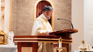 A man in a white and gold robe speaks at a wooden lectern in a church, with lit candles and a brick wall backdrop.