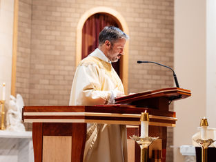 A man in a white and gold robe speaks at a wooden lectern in a church, with lit candles and a brick wall backdrop.