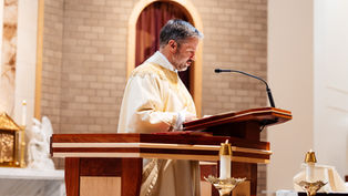 A man in a white and gold robe speaks at a wooden lectern in a church, with lit candles and a brick wall backdrop.