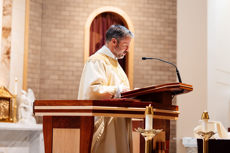 A man in a white and gold robe speaks at a wooden lectern in a church, with lit candles and a brick wall backdrop.