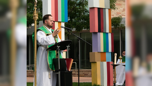 Priest in green vestments speaks into a microphone outdoors, with colorful pillar and greenery in the background. Calm and reflective mood.