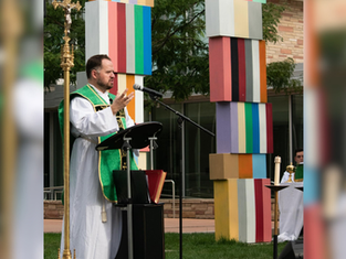 Priest in green vestments speaks into a microphone outdoors, with colorful pillar and greenery in the background. Calm and reflective mood.