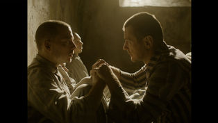 Two men in striped clothing sit in a dimly lit room, holding hands and gazing at each other intensely, evoking a somber mood.
