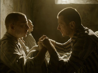 Two men in striped clothing sit in a dimly lit room, holding hands and gazing at each other intensely, evoking a somber mood.