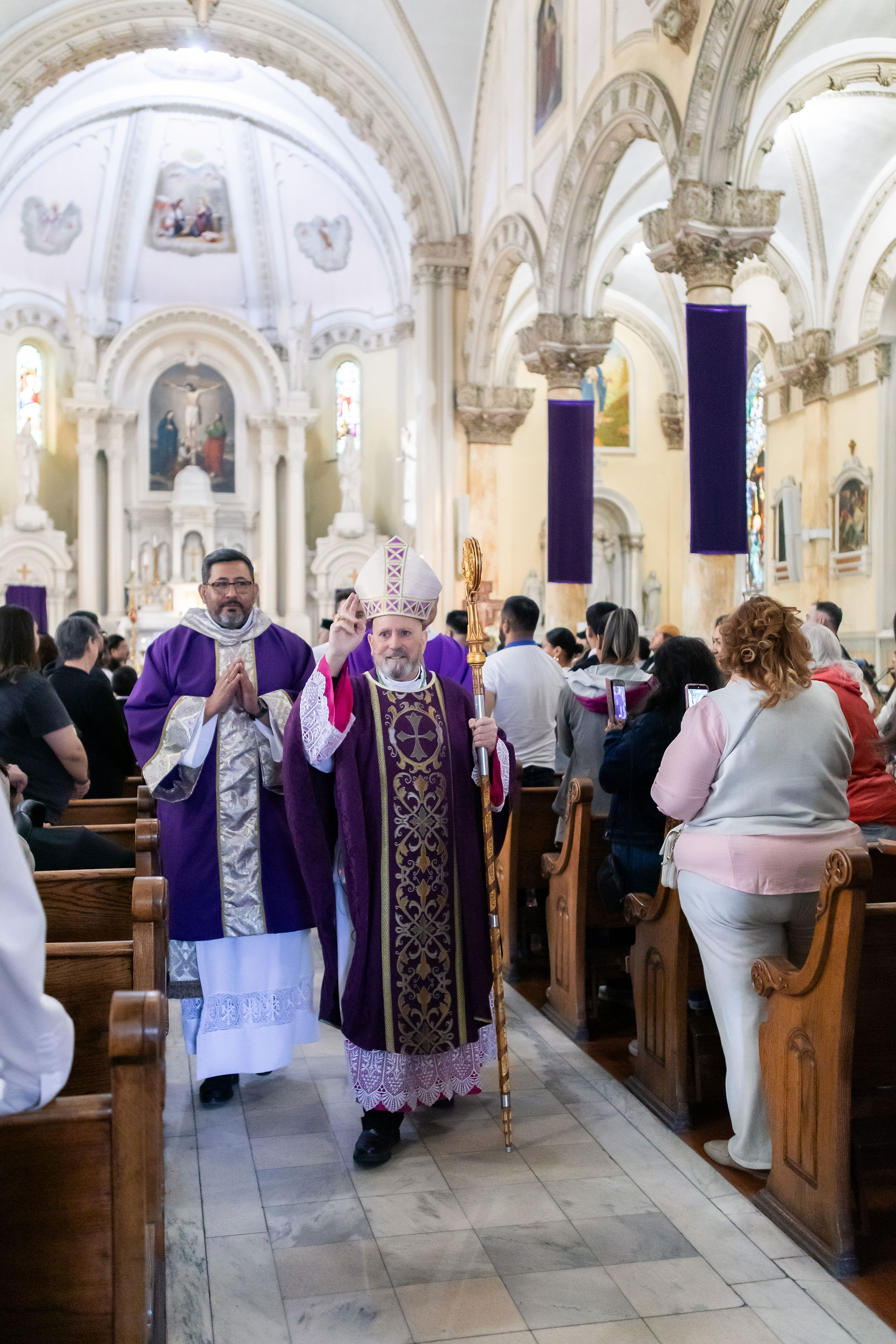 A bishop in ornate purple robes and miter walks through a crowded church, holding a staff. People watch, capturing the moment on phones.