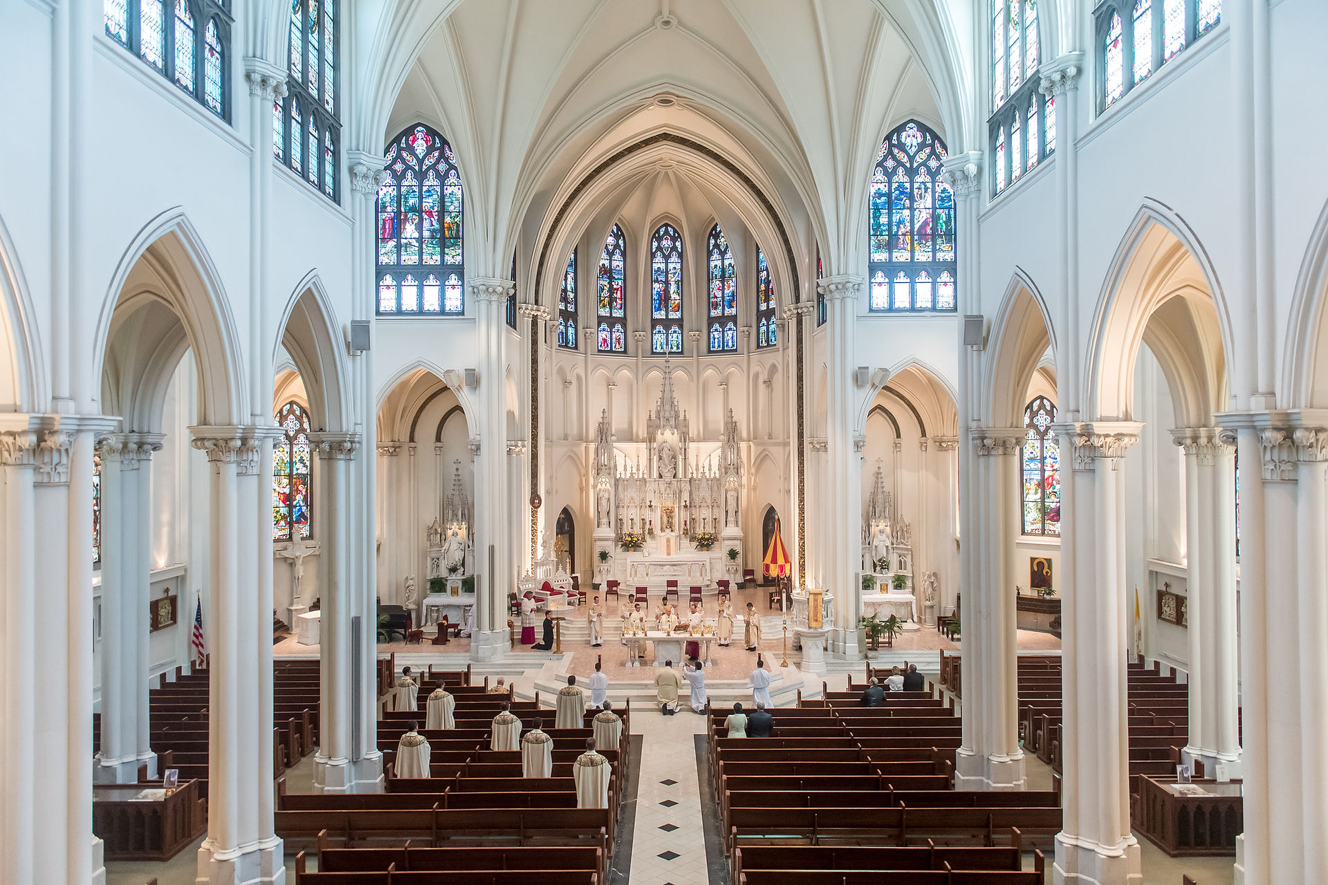 Interior of a grand cathedral with vaulted ceilings, stained glass windows, and clergy in white robes conducting a service. Peaceful ambiance.