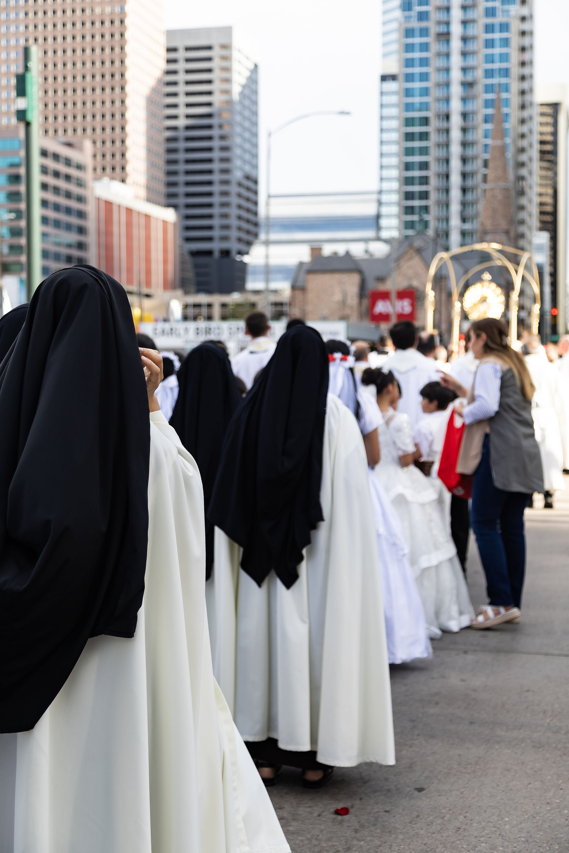 Nuns and children in white and black robes walk in a city street crowd near a religious procession, with tall buildings behind.