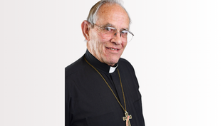 Elderly man in clerical attire with a cross necklace, smiling against a plain white background, conveying a warm, calm mood.