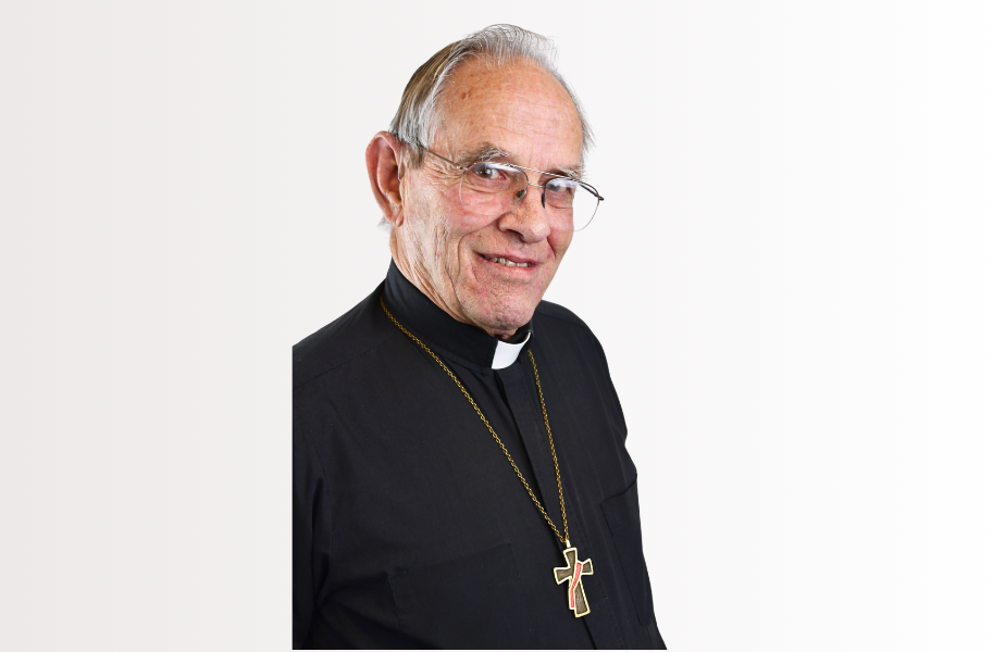 Elderly man in clerical attire with a cross necklace, smiling against a plain white background, conveying a warm, calm mood.