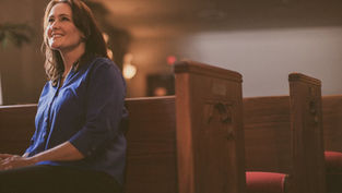 Woman in blue shirt sitting in a church pew, smiling softly. Warm lighting with wooden pews and blurred background create a serene mood.