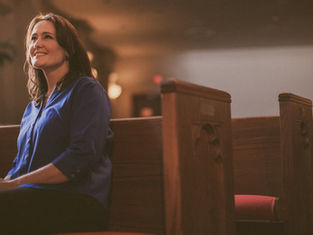 Woman in blue shirt sitting in a church pew, smiling softly. Warm lighting with wooden pews and blurred background create a serene mood.
