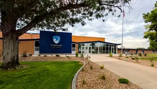 St. John Paul II High School building with blue sign, glass entrance, and American flag under a cloudy sky.