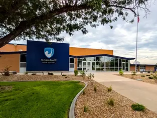 St. John Paul II High School building with blue sign, glass entrance, and American flag under a cloudy sky.