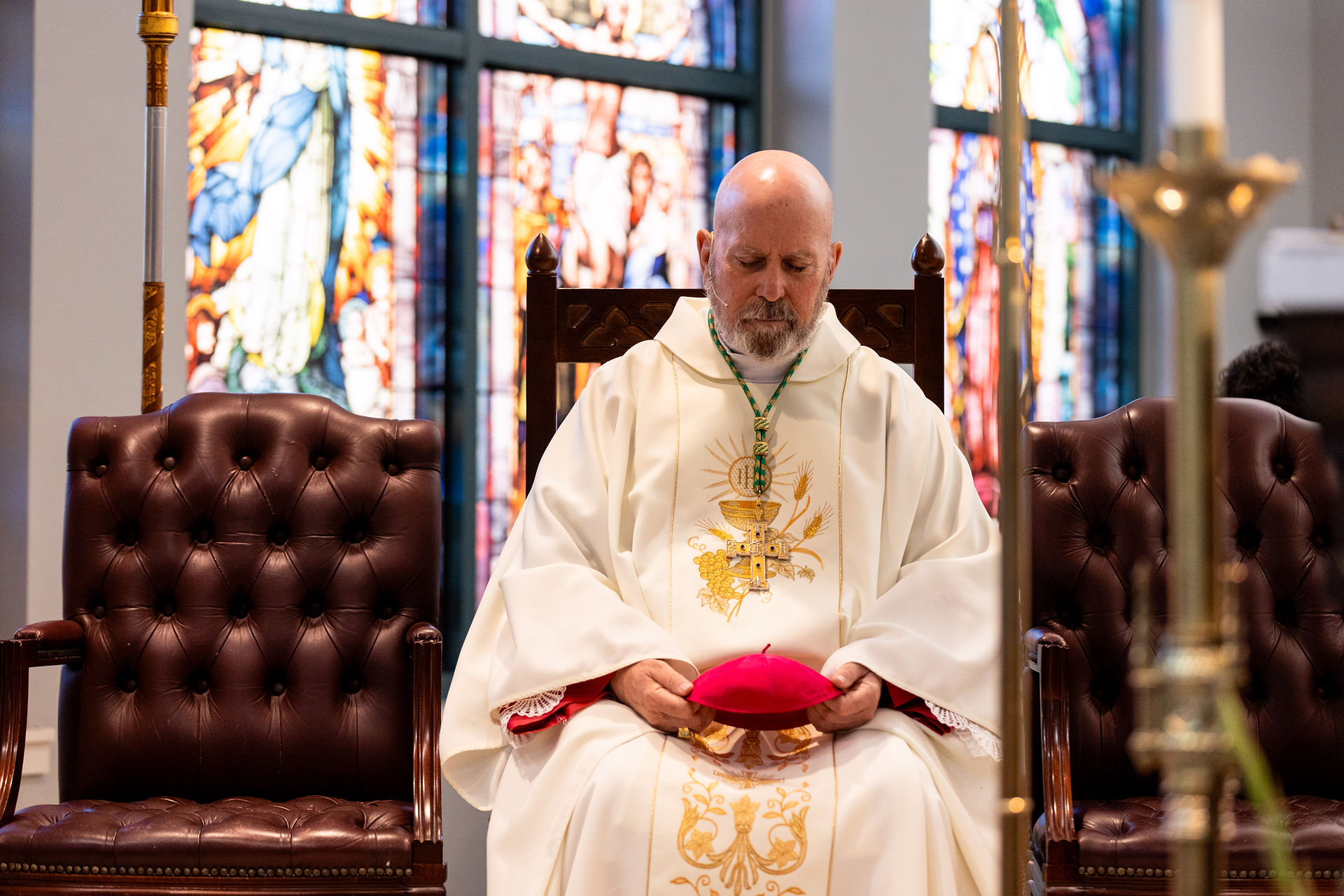 Seated clergy in ornate white robes holds a red cap, eyes closed in prayer. Stained glass windows create a colorful backdrop.
